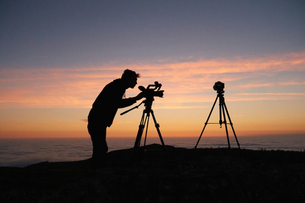 Photographe filmant un coucher de soleil avec une caméra sur trépied au sommet d'une montagne.