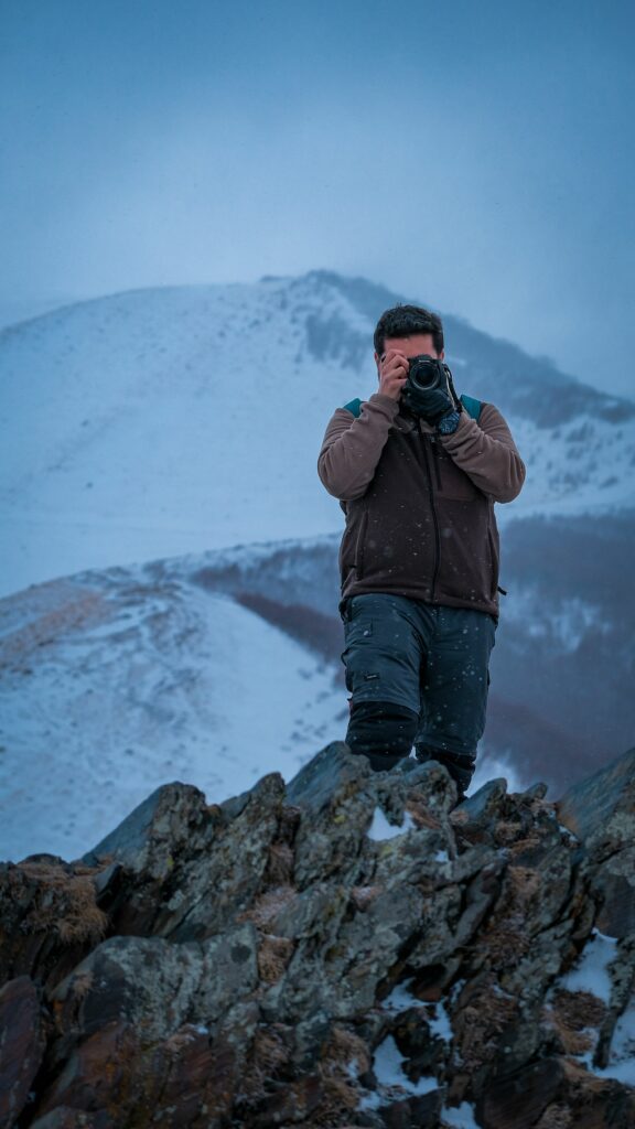 Photographe tenant un appareil photo dans un paysage de montagne enneigé sous un ciel couvert.