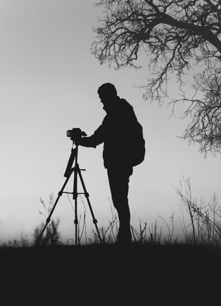 Silhouette d'un photographe réglant un appareil photo sur un trépied en extérieur, sous un arbre.